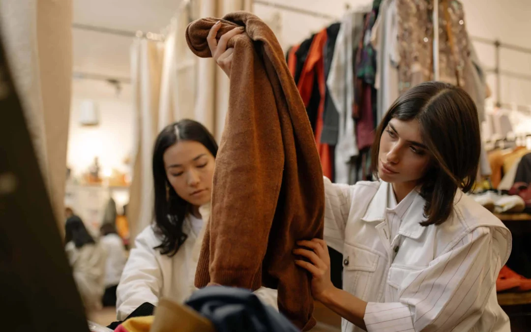 Two young women in a clothing shop examine a brown coat held up for inspection.