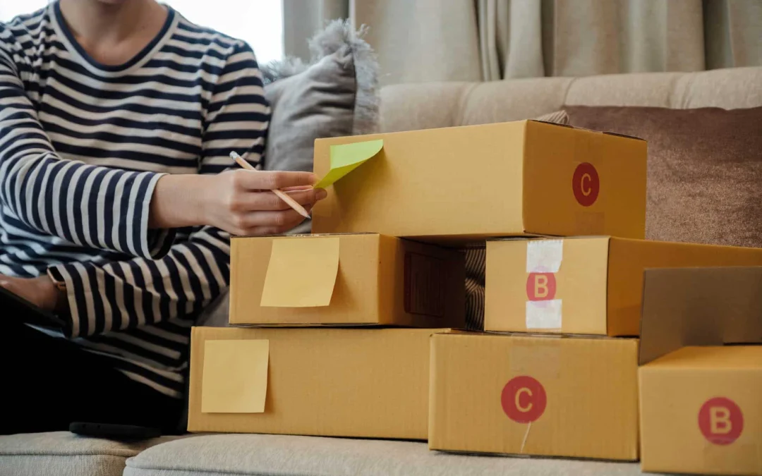 Person in a striped shirt writes on a sticky note while organizing boxes on a sofa in a living room.