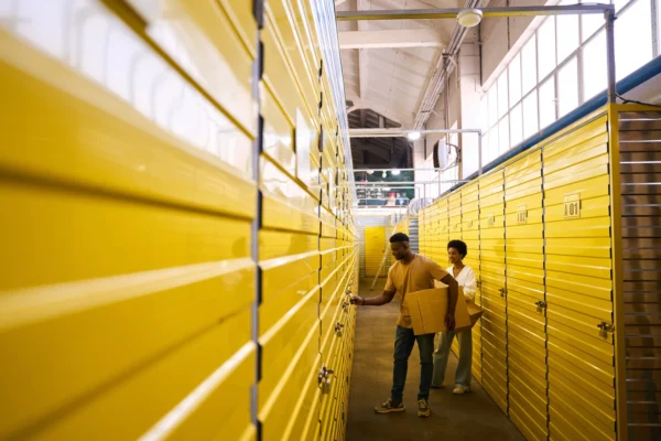 Two people move boxes down a narrow aisle of a self-storage facility with bright yellow unit doors, as the man opens a locker.