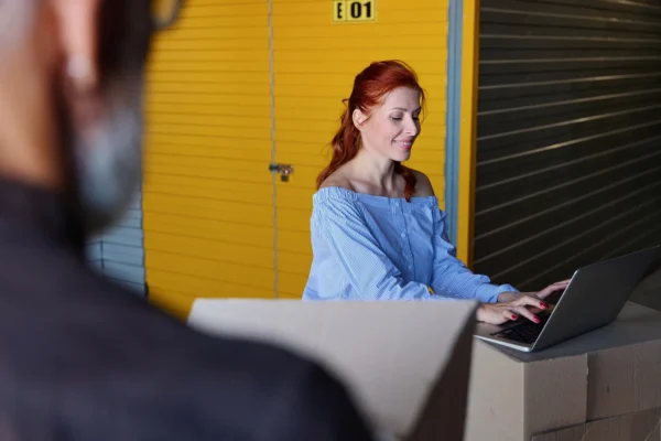 Woman with red hair in a blue off-shoulder striped top typing on a laptop in a warehouse with yellow shutters, boxes nearby.