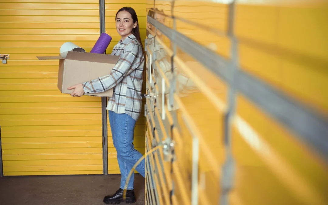 Woman in a plaid shirt holds a cardboard box with a purple yoga mat and foam roller, standing before yellow storage lockers.