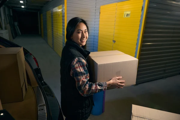 Smiling woman in a plaid shirt and black vest carries a large cardboard box beside an open car trunk in a storage facility with yellow storage doors behind her.