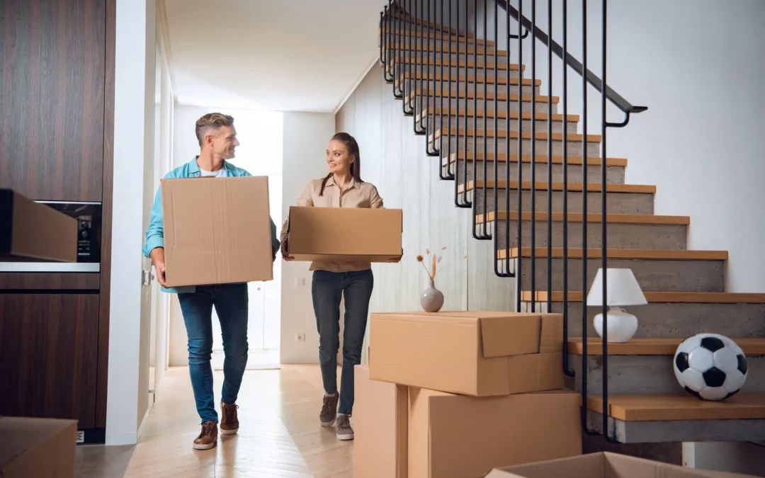 Couple carrying cardboard boxes in a modern home, stairwell to the right and packed boxes nearby.